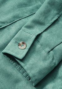 Gros plan sur une manche en tissu côtelé teal avec un unique bouton translucide gravé de "STONES" sur le poignet.