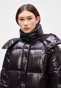 Geacă puf cu un finisaj lucios și matlasat, guler înalt, accente mari pe umeri și nasturi circulare. Include un patch cu logo.