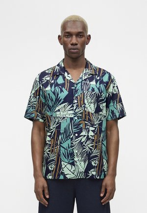 Modèle masculin aux cheveux blonds courts portant une chemise à manches courtes avec un motif abstrait de feuilles vertes et brunes.