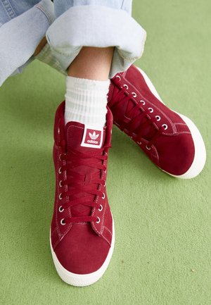 Personne portant un jean bleu clair retroussé, des chaussettes blanches et des baskets montantes rouges Adidas sur un sol recouvert d'un tapis vert.