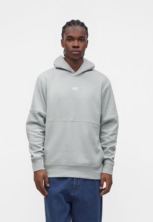 Junger Mann mit geflochtenem Haar trägt einen hellgrauen Nike-Hoodie und blaue Jeans, stehend vor einem schlichten weißen Hintergrund.