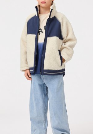 Ragazzo con giacca in pile crema e blu navy con cerniera, maglietta con grafica e jeans larghi azzurri, in piedi con una mano in tasca.