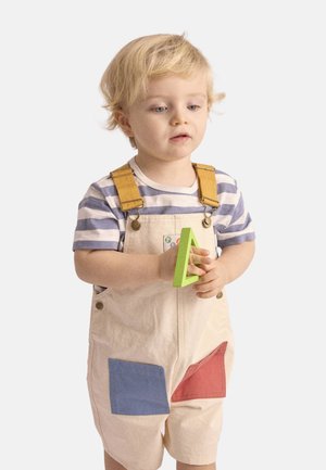 Jeune enfant en chemise rayée et salopette avec poche en patch tenant un jouet triangulaire en plastique vert, regardant légèrement vers le bas.