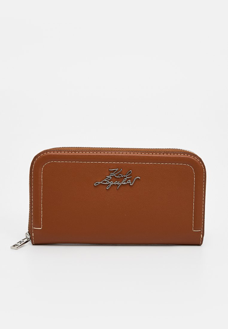 Portefeuille marron en simili cuir avec une fermeture zippée, des coutures blanches et un accent métallique avec la signature sur le devant. Forme rectangulaire, texture lisse.
