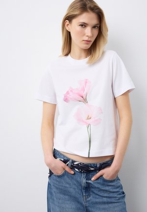 Mujer joven con cabello rubio, vestida con camiseta blanca con estampado de flores rosas y jeans azules con cinturón oscuro, de pie contra una pared blanca.