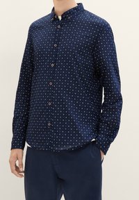 Marineblaues Langarmhemd mit Button-Down-Kragen, versehen mit einem weißen Sternenmuster und braunen Knöpfen. Weiche Textur, taillierte Passform.