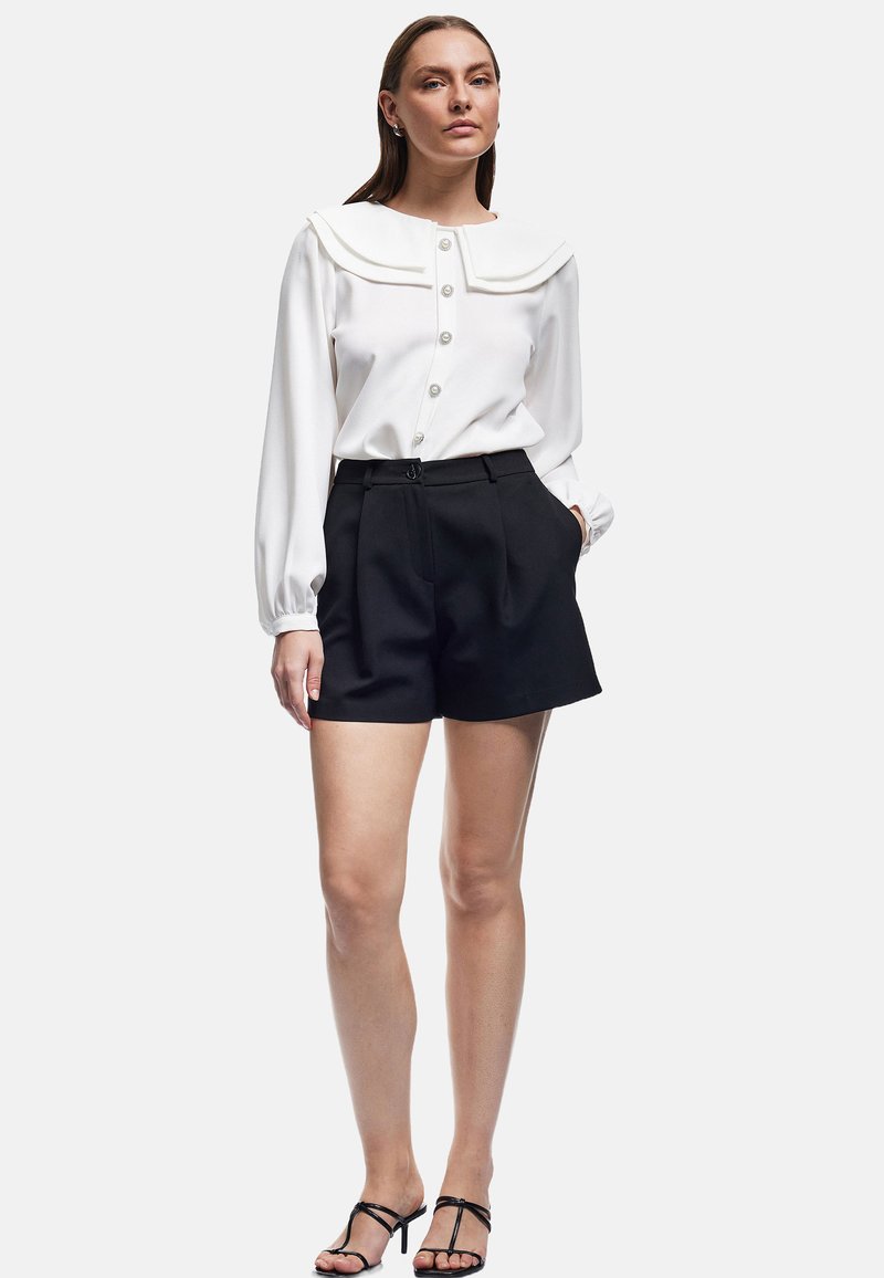 Kvinna som står med händerna i svarta skräddarsydda shorts, vit blus med bred krage och pärlknappar, iförd svarta högklackade sandaler.
