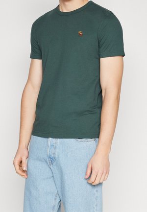 Zielona bawełniana koszulka z krótkimi rękawami, okrągłym dekoltem i haftowanym wzorem na piersi. W zestawie z jasnoniebieskimi dżinsami.