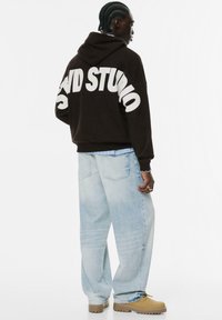 Mand med fletninger, iført en sort hættetr�øje med teksten "STUDIO", lyseblå baggy jeans og brune støvler, står i et minimalistisk hvidt rum.