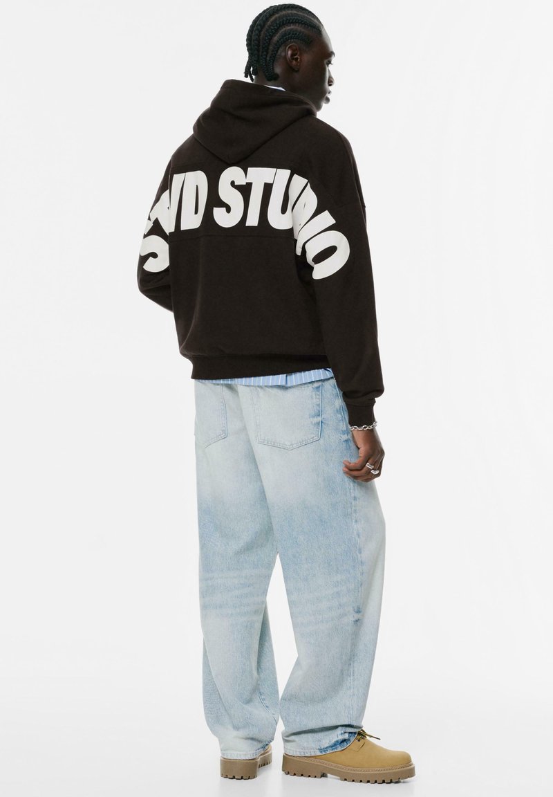 Mand med fletninger, iført en sort hættetrøje med teksten "STUDIO", lyseblå baggy jeans og brune støvler, står i et minimalistisk hvidt rum.