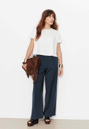 Femme portant un haut blanc à manches courtes, un pantalon large à fines rayures bleu marine, des sandales compensées noires, tenant un sac à main marron à franges, debout devant un mur blanc.