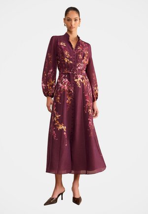 Femme portant une robe midi violette à manches longues avec motifs floraux, boutons sur le devant, taille ceinturée et talons pointus marron sur fond blanc.
