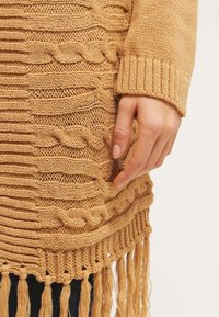 Musztardowy dzianinowy sweter z fakturowanym wzorem warkocza i długim, frędzlowanym dołem. Posiada ściągacze na mankietach, a na materiale spoczywa ręka.