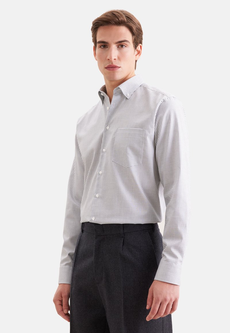 Jeune homme portant une chemise à manches longues blanche et grise à carreaux rentrée dans un pantalon gris foncé, debout devant un fond uni.