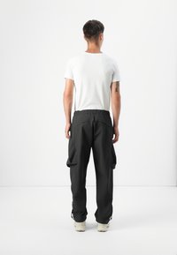 En person står med ryggen til og bærer en tætsiddende hvid T-shirt og løse sorte cargo-bukser, mod en minimalistisch hvid baggrund.