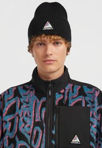 Berretto a coste nero e giacca in pile colorata con motivo geometrico in blu, rosa e nero, dotata di chiusura con zip e tasca sul petto.