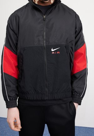 Personne portant une veste noire Nike Air avec des panneaux rouges sur les manches et une fermeture éclair à l'avant, debout avec une main dans la poche.