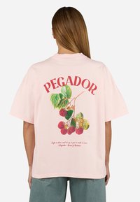 Šviesiai rožinė perteklinė T-marškiniai su „PEGADOR“ tekstu, aviečių grafiku ir citata, atspausdinta arti apvado. Kasdienė medvilnės medžiaga.