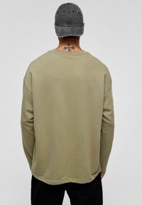 Olijfgroene long-sleeve shirt met een ronde zoom en een relaxte pasvorm, in combinatie met een donkergrijze pet. De textuur lijkt zacht en glad.