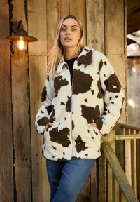 Witte fleece zip-up jas met bruine koeienprint, voorzien van een kraag en voorzakken, gedragen over een zwarte top met blauwe spijkerbroeken.
