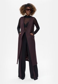 Kobieta z kręconymi włosami nosząca duże okulary przeciwsłoneczne, duże kolczyki, przezroczystą czarną bluzkę, długą ciemną kamizelkę i szerokie spodnie, z rękami w kieszeniach.