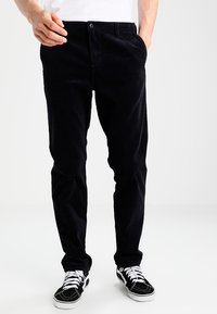 Homme portant un pantalon en velours côtelé noir, des baskets noir et blanc, et une chemise blanche, se tenant avec une main dans la poche.