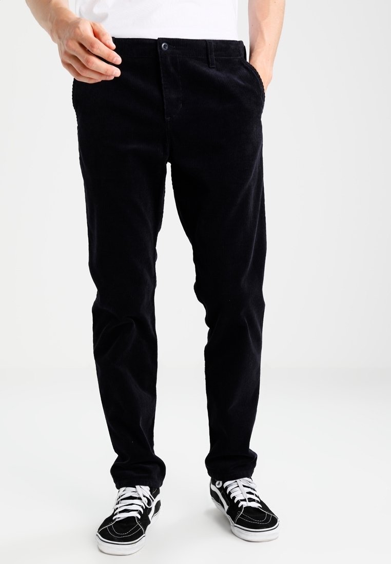 Homme portant un pantalon en velours côtelé noir, des baskets noir et blanc, et une chemise blanche, se tenant avec une main dans la poche.