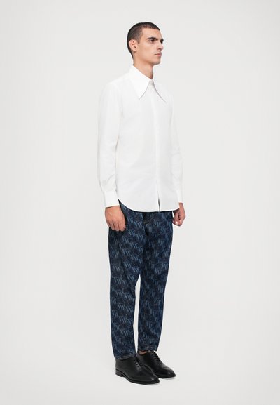 Camisa blanca de manga larga con un gran cuello, combinada con pantalones azules con un patrón que presenta un diseño gráfico repetido, y zapatos negros.