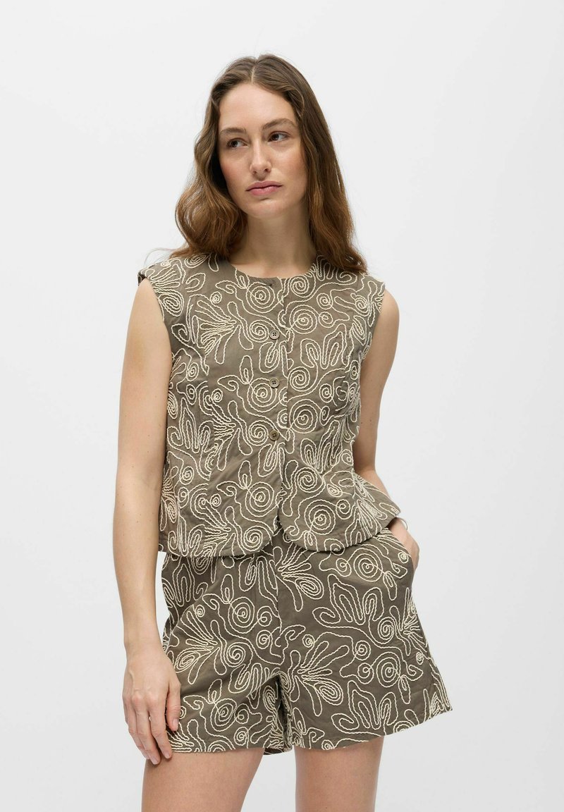 Femme aux longs cheveux bruns portant un haut sans manches assorti et un short avec une broderie abstraite en forme de tourbillon beige sur tissu olive.