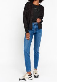 Svart, glittrig texturerad cardigan över en svart t-shirt med tryck, kombinerad med blå raka jeans och crema- och svart sneakers.