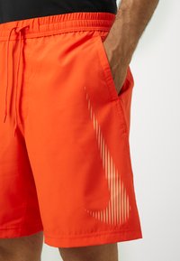 Shorts de bain Nike orange avec un design de logo texturé sur le côté gauche, taille élastique et poches latérales. Texture de tissu lisse.