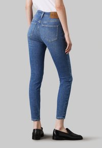 Jeans de mezclilla azul ajustados con una cintura alta, cinco bolsillos y un parche de logo de cuero marrón en la parte posterior. Textura suave con costuras sutiles.