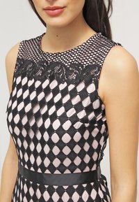 Czarna sukienka z koronką i wzorem w diamenty, z wysokim dekoltem i czarnym satynowym paskiem w talii. Góra jest półprzezroczysta.