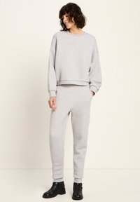 Frau in hellgrauem Sweatshirt und passender Jogginghose mit schwarzen Stiefeletten, die vor einem schlichten Hintergrund steht, die Hände in den Taschen und lächelt.