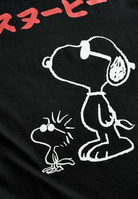 Sort stof med hvide tegneseriefigurer som Snoopy og Woodstock. Tekst ovenfor i fed rød japansk skrift. Enkel, legesyg design.