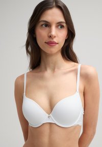 Witte beha met gladde voorgevormde cups, verstelbare bandjes en kanten accenten langs de zijkanten. Bevat een klein centraal decoratief detail.
