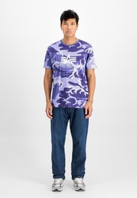 Ljubičasta vojaška majica s kratkimi rokavi, ki ima belo logo. V kombinaciji s temno modrimi kavbojkami in sivimi športnimi čevlji.