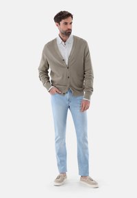 Hellbrauner Cardigan, geknöpft, über einem hellgrauen Hemd getragen, kombiniert mit hellblauen Jeans und beigen Slippern, vor einem weißen Hintergrund.