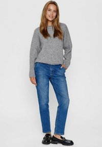 Grijze geribbelde trui, relaxed fit, gecombineerd met blauwe denim jeans. Zwarte instappers met gouden hardware maken de outfit compleet. Eenvoudige achtergrond.