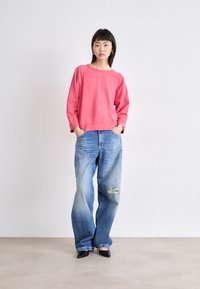 Sudadera rosa con mangas raglán combinada con unos vaqueros anchos azules con una rodilla desgastada. Unos zapatos negros de punta completan el conjunto.