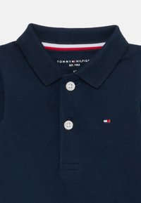 Polo de culoare albastru marin, fabricat din bumbac, cu un finisaj texturat, dotat cu o plachetă cu trei nasturi și un mic logo brodat pe piept.
