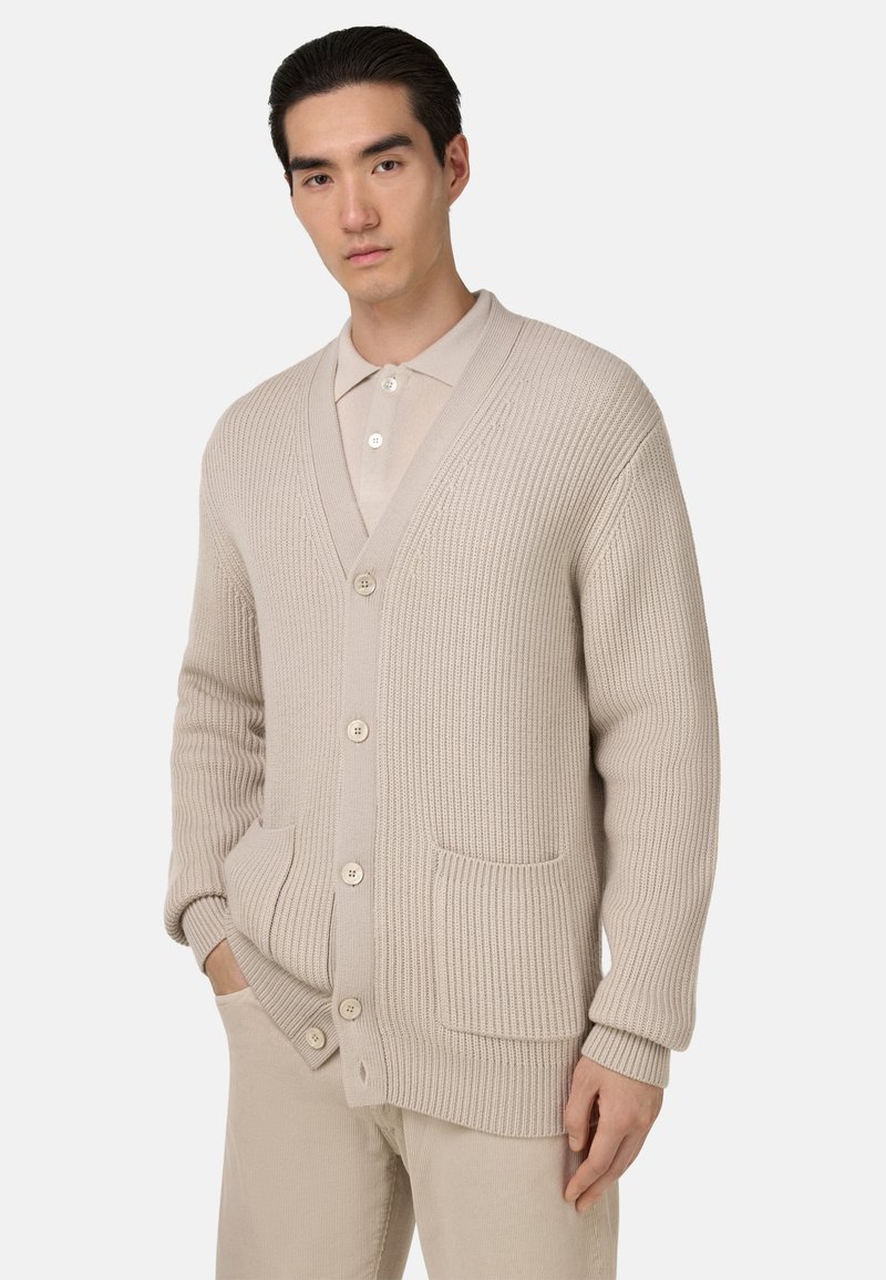 Beżowy kardigan w prążki z dekoltem w serek, trzema guzikami, dwiema przednimi kieszeniami i teksturowanym wzorem dzianiny, noszony na beżowej koszuli z kołnierzykiem.