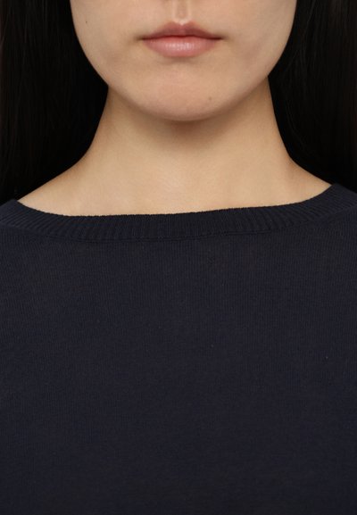 Jūras zila džemperis ar ribotu kuģa kakla apdari un mīkstu tekstūru. Audums izskatās viegls ar smalku spīdumu, uzsverot vienkāršību.