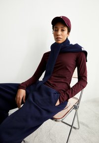 Bordeaux-rotes Langarmoberteil, marineblauer Schal, der über die Schultern drapiert ist, marineblaue Jogginghose und eine bordeaux-rote Mütze, sitzend auf einem Stuhl mit chromrahmen.