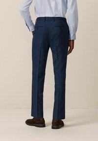 Pantalones marinos con un corte ajustado, que presentan dos bolsillos traseros con cierres de botón y una tela lisa y texturada. Zapatos marrones complementan el conjunto.