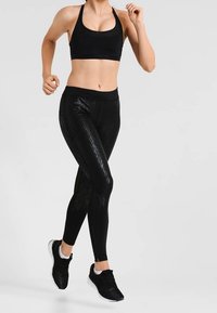 Il reggiseno sportivo nero e i leggings presentano una texture lucida con strisce verticali. Il modello indossa scarpe da corsa nere e l'outfit è aderente per facilitare i movimenti.