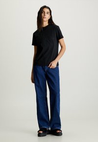 T-shirt in cotone nero con logo impresso, abbinato a jeans in denim blu a gamba larga. Il modello indossa sandali neri chunky. Design minimalista.