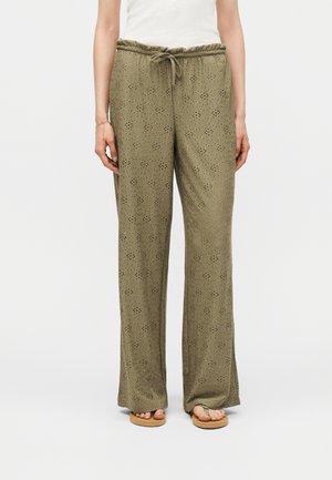 Γυναίκα που φοράει ελαιδί φαρδιά παντελόνια με λουλουδάτο μοτίβο με οπές και ζωνάκι στη μέση, συνδυασμένα με μπεζ σανδάλια και λευκό τοπ.