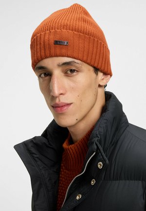 Beanie a coste arancione con etichetta nera, indossato con un giubbotto imbottito scuro e un maglione arancione a trama, su sfondo bianco.