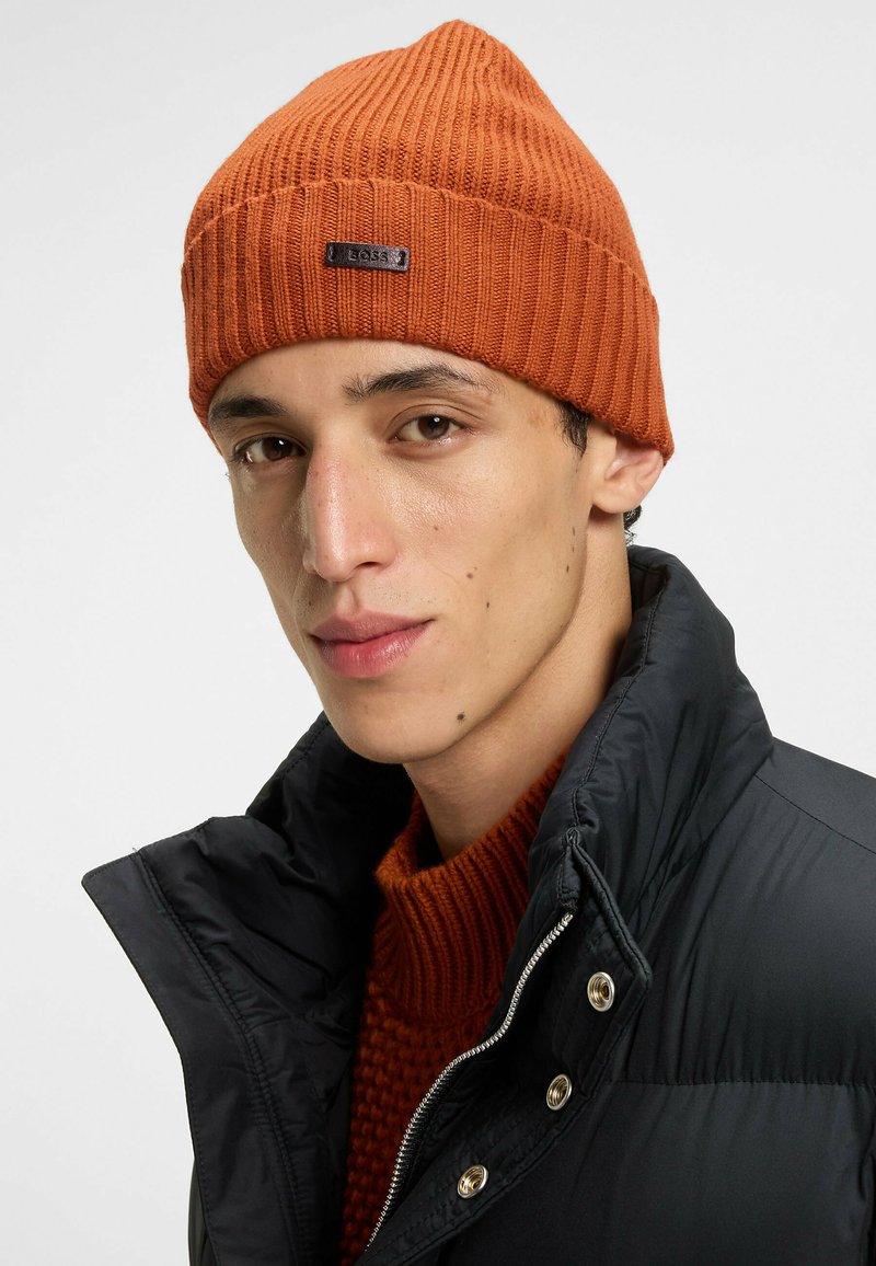 Een oranje geribbelde gebreide beanie met een zwart logo etiqueta, gedragen met een donkere gewatteerde jas en een gestructureerd oranje sweatshirt, tegen een witte achtergrond.